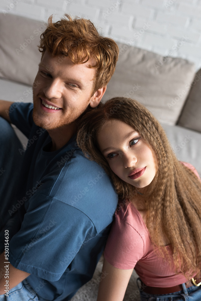 teenage girl leaning on back of happy boyfriend with red hair in living room. teenage girl leaning on back of happy boyfriend with red hair in living room.