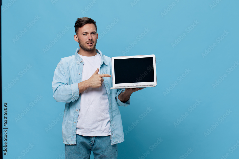 Smiling happy tanned handsome man in casual basic t-shirt point finger at new purchase laptop posing isolated on blue studio background. Copy space Banner Mockup. Electronics repair IT concept Smiling happy tanned handsome man in casual basic t-shirt point finger at new purchase laptop posing isolated on blue studio background. Copy space Banner Mockup. Electronics repair IT concept