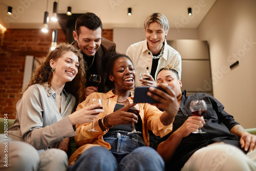 Young cheerful black woman taking selfie with group of intercultural friends gathered in living room for home party on weekend