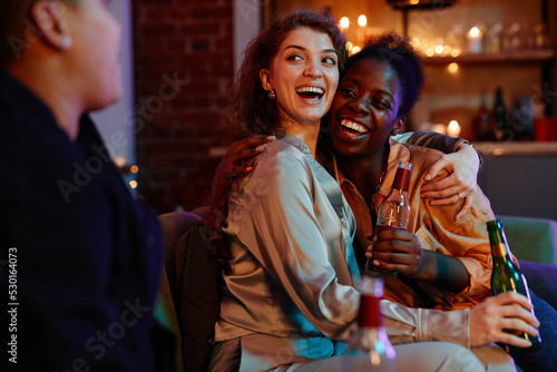 Young laughing woman with bottle of beer embracing her girlfriend sitting next to her on couch and looking at another girl