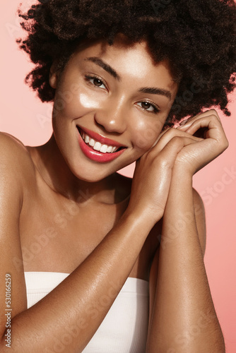 Beautiful smiling african american woman, teen skin care cosmetics advertisement. Portrait of girl with hydrated face, using nourishing cream after shower, pink background
