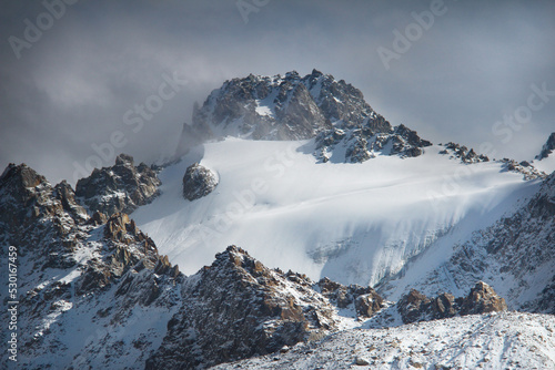 A large rocky mountain with a rocky peak  on the mountain the Tuyuk-Su glacier is covered with deep snow  the sun is shining on the right  the sky is with dark thick clouds