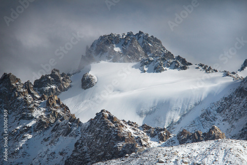 A large rocky mountain with a rocky peak, on the mountain the Tuyuk-Su glacier is covered with deep snow, the sun is shining on the right, the sky is with dark thick clouds