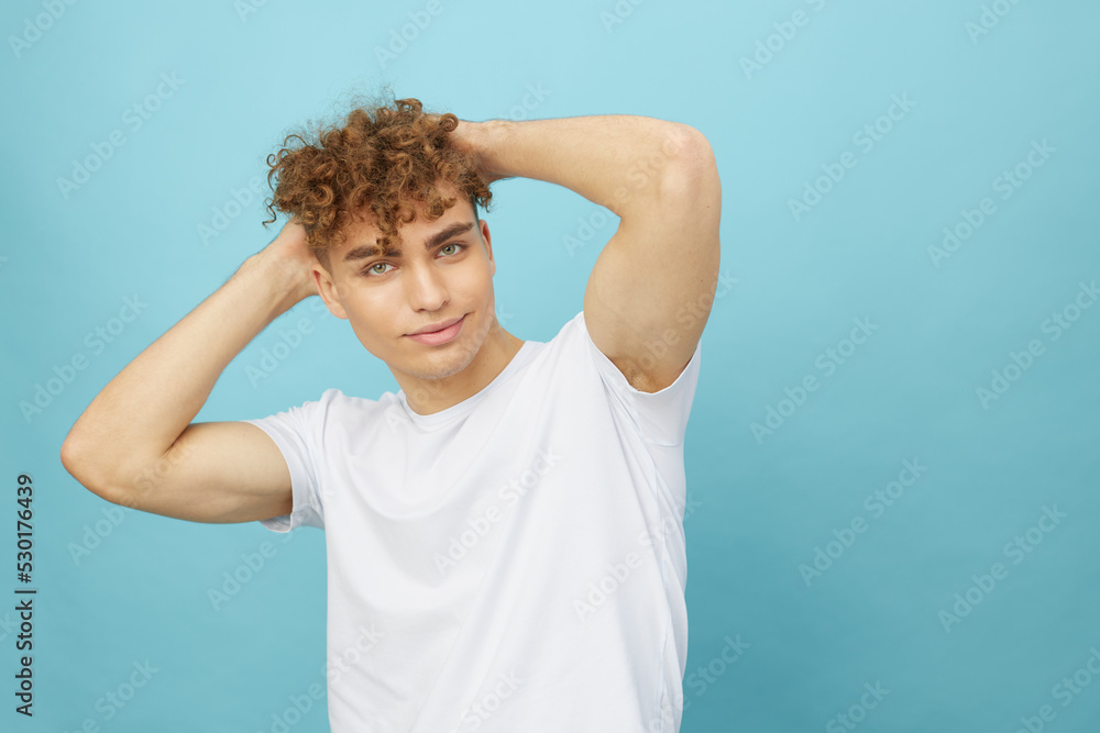 A young man on an isolated blue wall with his mouth wide open from positive emotions holds his hand in curly hair A young man on an isolated blue wall with his mouth wide open from positive emotions holds his hand in curly hair