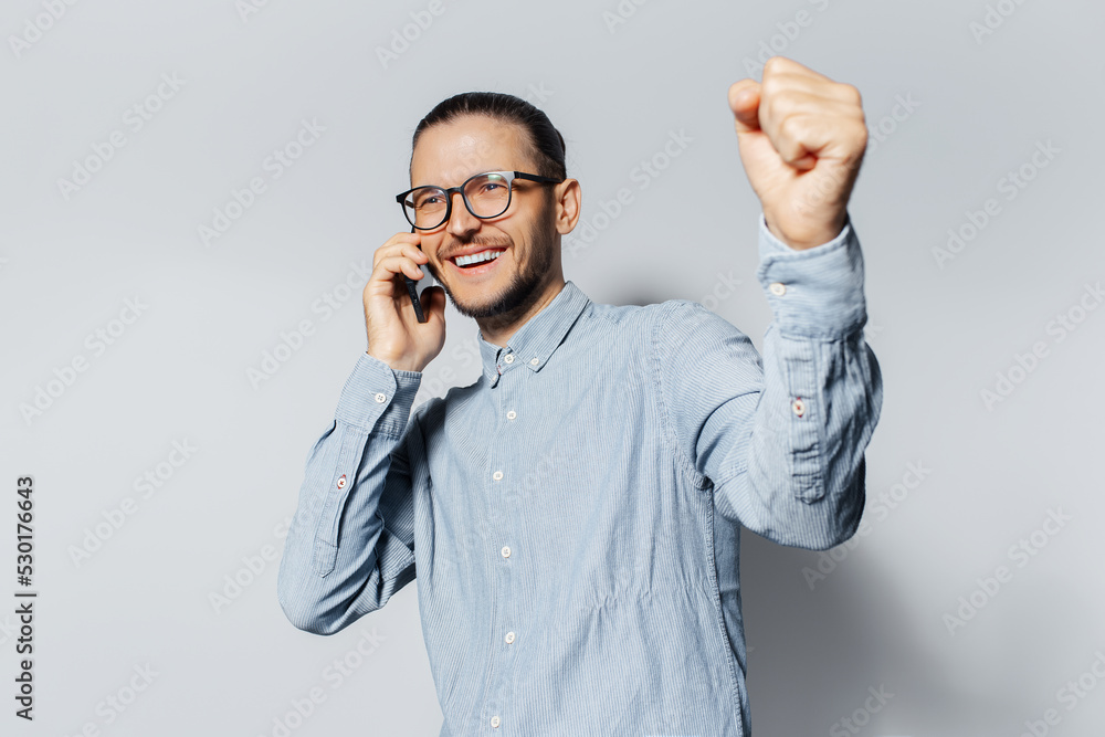 Yes. Studio portrait of young happiness guy talking on smartphone, doing winning gesture on white background. Wearing eyeglasses and blue shirt. Yes. Studio portrait of young happiness guy talking on smartphone, doing winning gesture on white background. Wearing eyeglasses and blue shirt.