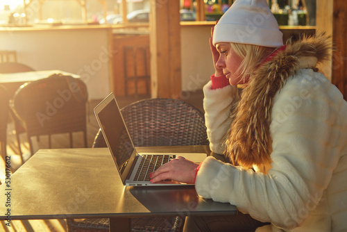 A woman in a sweater and a fur coat is sitting on the terrace of a cafe with a laptop. 