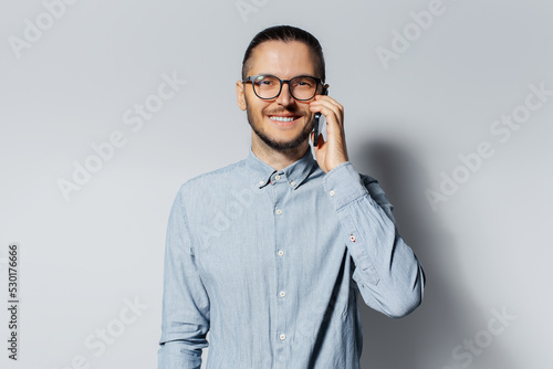 Studio portrait of young smiling man talking on smartphone, wearing eyeglasses and blue shirt on grey background.