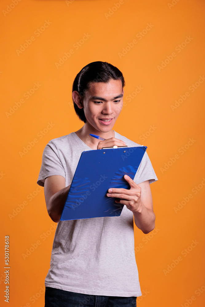 Smiling asian man taking notes in blue clipboard, student writing in notepad with pen. Person filling survey form, corporate plan, office worker holding documents, medium shot Smiling asian man taking notes in blue clipboard, student writing in notepad with pen. Person filling survey form, corporate plan, office worker holding documents, medium shot