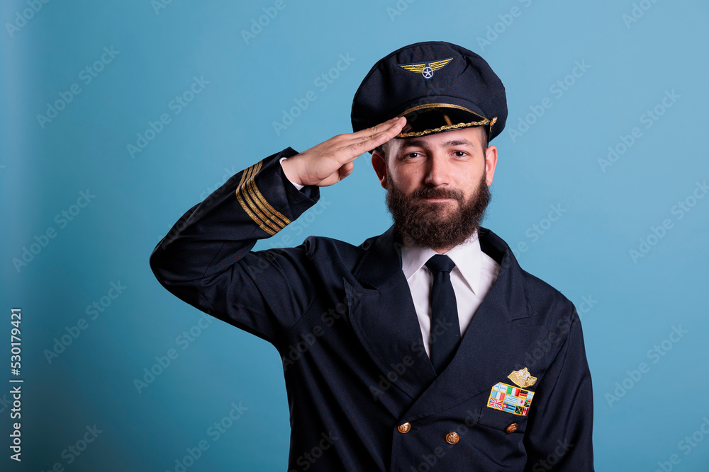 Confident airplane pilot saluting, wearing uniform and hat front view portrait, plane captain looking at camera. Civil aviator with airline wings badge on jacket, professional aircrew member Confident airplane pilot saluting, wearing uniform and hat front view portrait, plane captain looking at camera. Civil aviator with airline wings badge on jacket, professional aircrew member