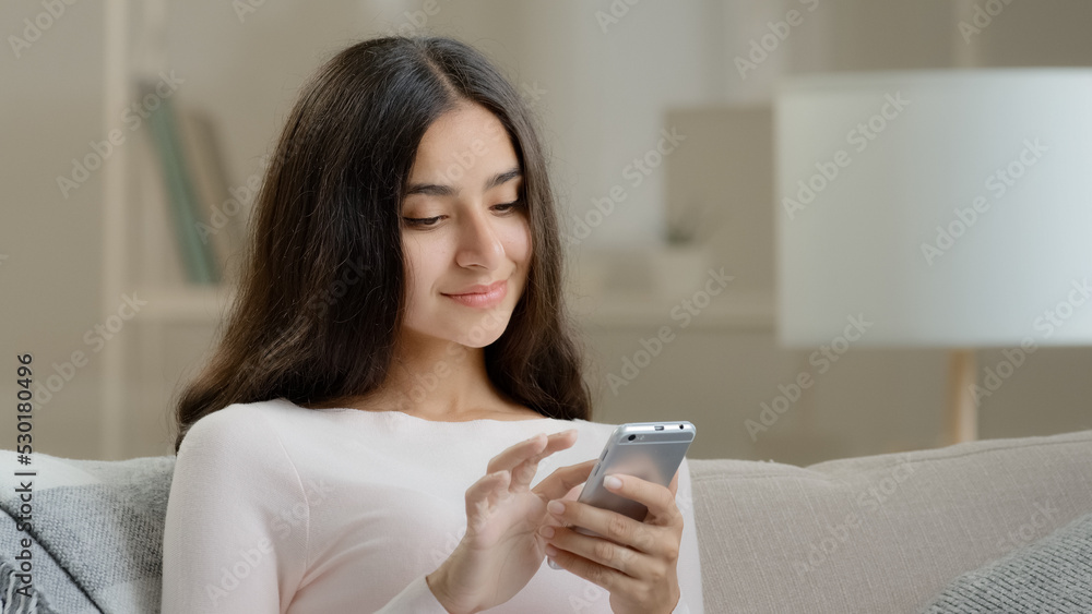 Female young user freelancer calm attractive girl arab hispanic woman sits on sofa at home scrolls inform pages on telephone device chatting makes internet purchases shopping online in clothing store Female young user freelancer calm attractive girl arab hispanic woman sits on sofa at home scrolls inform pages on telephone device chatting makes internet purchases shopping online in clothing store