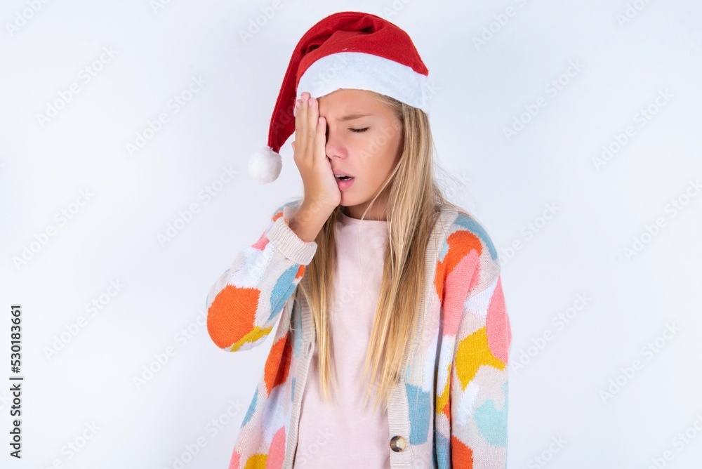 little kid girl with Christmas hat wearing yarn jacket over white background Yawning tired covering half face, eye and mouth with hand. Face hurts in pain. little kid girl with Christmas hat wearing yarn jacket over white background Yawning tired covering half face, eye and mouth with hand. Face hurts in pain.