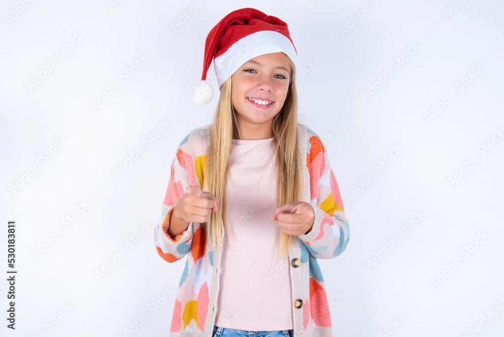 little kid girl with Christmas hat wearing yarn jacket over white background pointing fingers to camera with happy and funny face. Good energy and vibes. little kid girl with Christmas hat wearing yarn jacket over white background pointing fingers to camera with happy and funny face. Good energy and vibes.