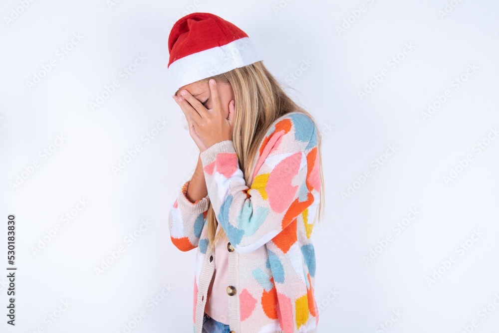Sad little kid girl with Christmas hat wearing yarn jacket over white background crying covering her face with her hands. Sad little kid girl with Christmas hat wearing yarn jacket over white background crying covering her face with her hands.