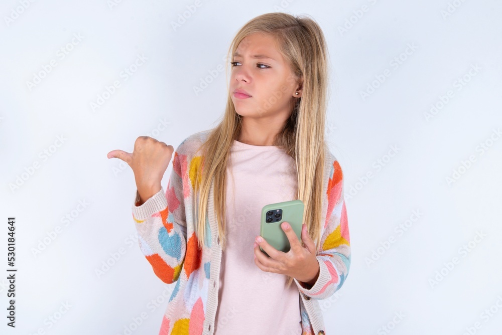 little kid girl wearing colorful yarn jacket over white background points thumb away and shows blank space aside, holds mobile phone for sending text messages. little kid girl wearing colorful yarn jacket over white background points thumb away and shows blank space aside, holds mobile phone for sending text messages.