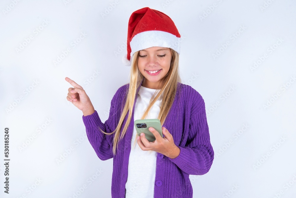 Smiling little kid girl with Christmas hat wearing yarn jacket over white background pointing finger at blank space holding phone in one hand