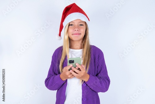 Portrait of serious confident little kid girl with Christmas hat wearing yarn jacket over white background holding phone in two hands