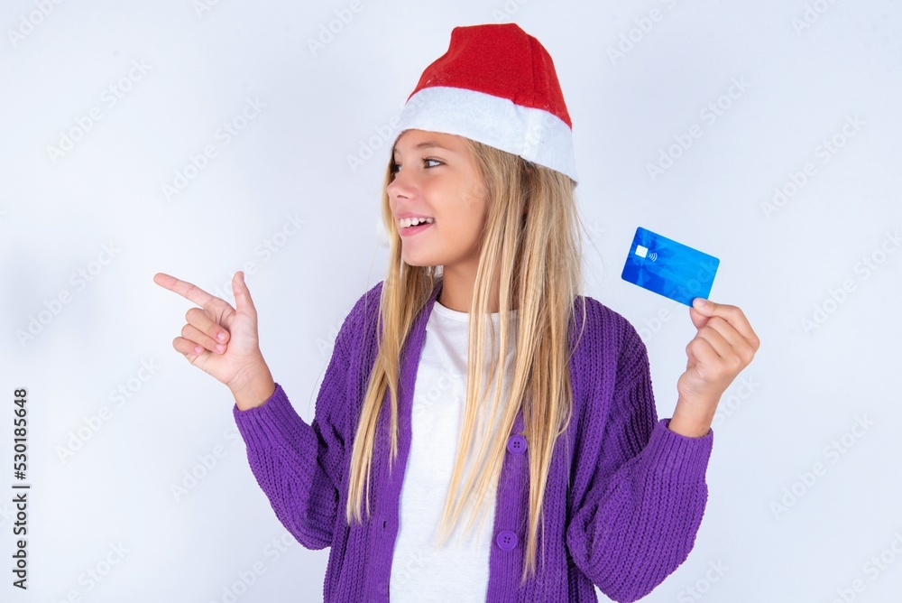 Curious smiling little kid girl with Christmas hat wearing yarn jacket over white background showing plastic bank showing finger copyspace Curious smiling little kid girl with Christmas hat wearing yarn jacket over white background showing plastic bank showing finger copyspace