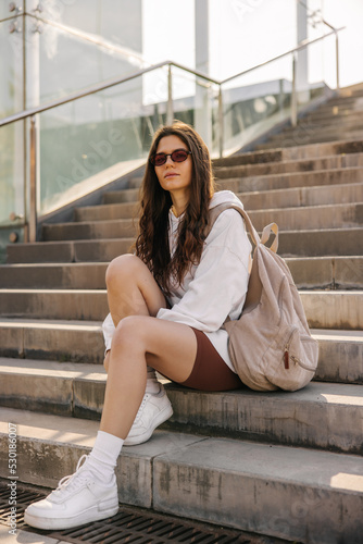 Full-length view of fair-skinned young woman sitting on stairs in sunglasses. Look with smile to camera  holding backpack at street. Rest time concept 