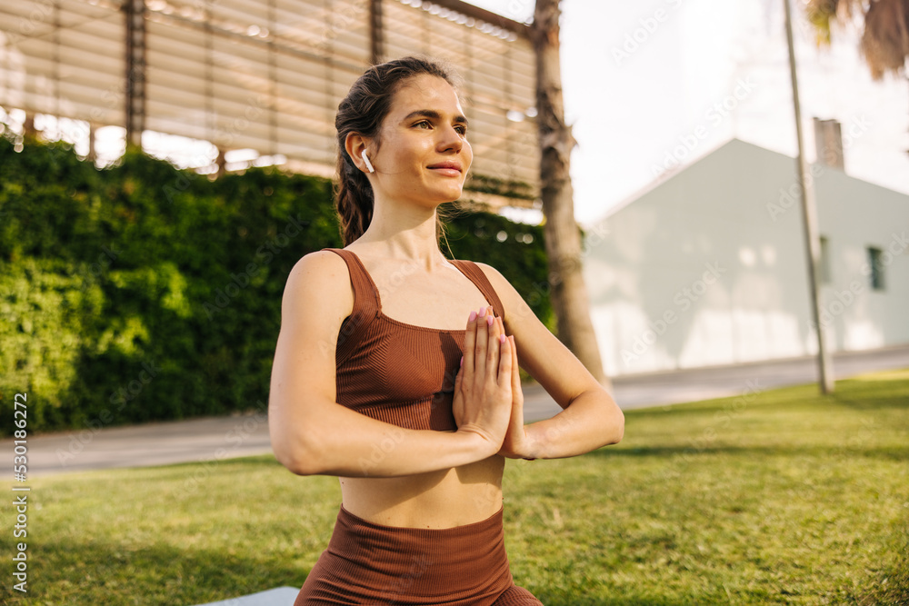 Beautiful pretty woman looking away, in yoga pose sitting at park and smiling. Keep hands together and listening meditation on headphones. Use technology, rest time concept 