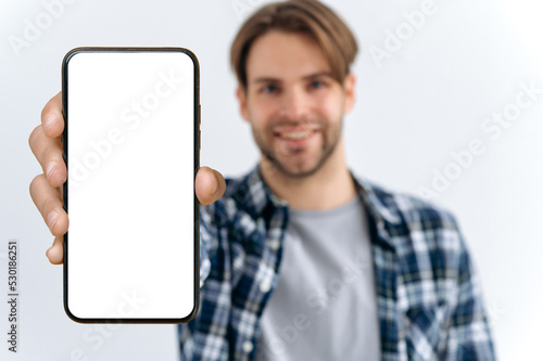 Defocused photo of a caucasian young man, looks at camera, hold in hand smartphone with white blank mock-up screen for presentation or advertising, stand on isolated white background, smiles