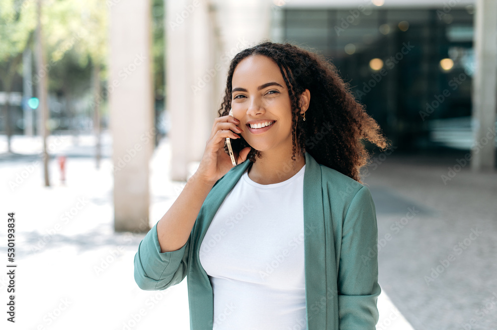 Wireless communication. Busy positive latino or brazilian woman, successful realtor, business lady in elegant clothes, has a pleasant conversation on a cell phone while standing outdoors, smiling Wireless communication. Busy positive latino or brazilian woman, successful realtor, business lady in elegant clothes, has a pleasant conversation on a cell phone while standing outdoors, smiling