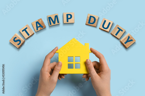 Woman holding house figure and wooden cubes with text Stamp Duty on light blue background, top view photo