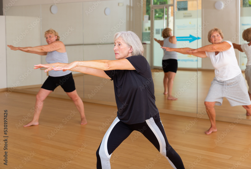 Portrait of athletic european aged woman doing stretching warm-up during group workout at gym .