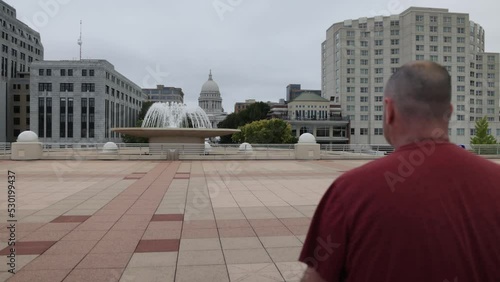 Caucasian male in maroon shirt walking to camera on the Monona Terrace in Madison, Wisconsin with gimbal video behind. photo