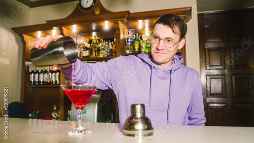 Man bartender with cocktail shaker and glass at bar. Male bartender from a shaker pours a red cocktail into a glass on bar counter.