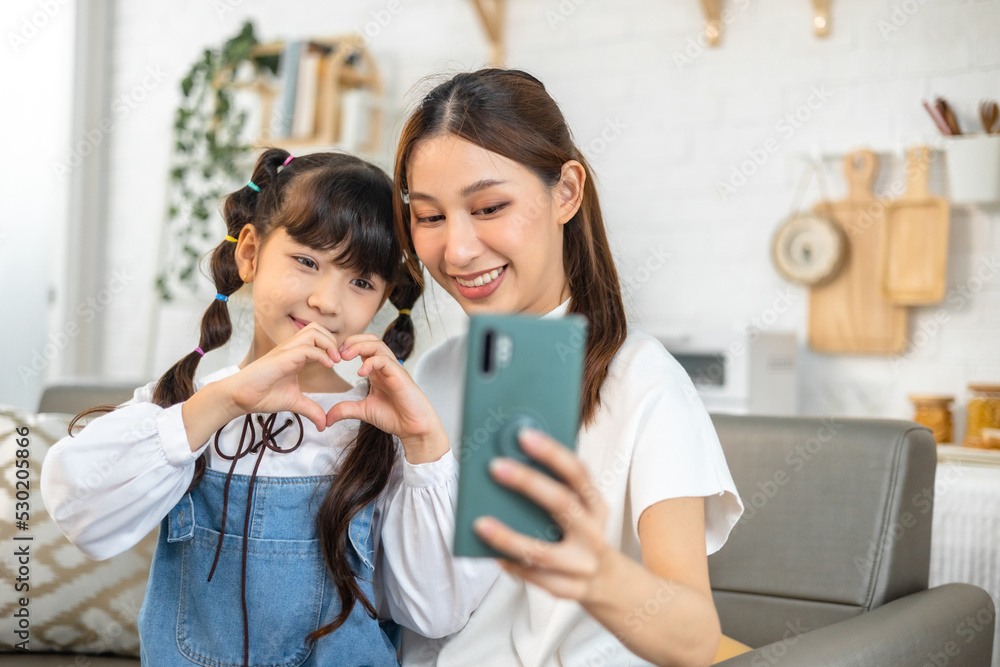 Young asian mother and her daughter are making selfie using a phone, hugging and smiling while sitting on the sofa at home. Young asian mother and her daughter are making selfie using a phone, hugging and smiling while sitting on the sofa at home.