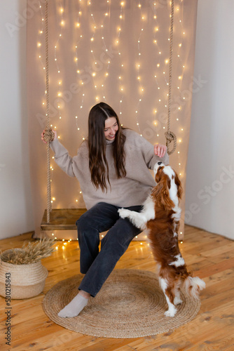 Woman with King Charles spaniel dog sitting at swing in festively decorated house.