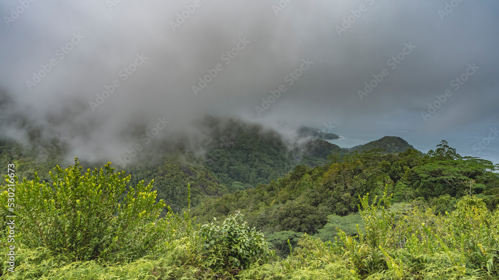 Low rain clouds and fog hung over the mountains. The valley has lush green tropical vegetation. The turquoise ocean is visible in the distance. Seychelles. Mahe Island