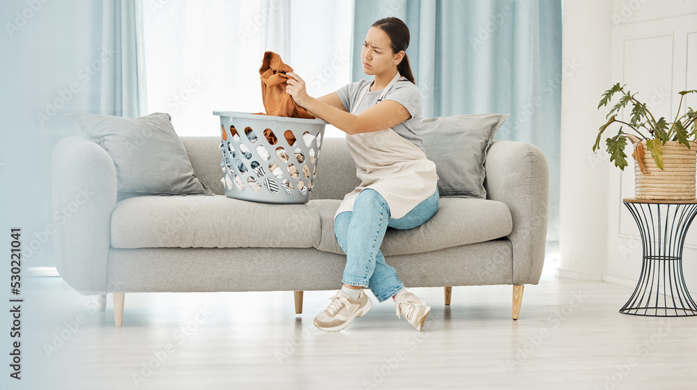Asian woman, laundry or clothes basket on sofa in house living room and looking confused at dirty stains. Stress, anxiety or worry of person with washing clothing or cleaning fabric in home interior
