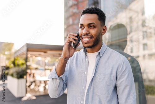 Modern african young guy with beard walks around city and calls on mobile phone. Dark-haired man wears blue shirt. Communication concept