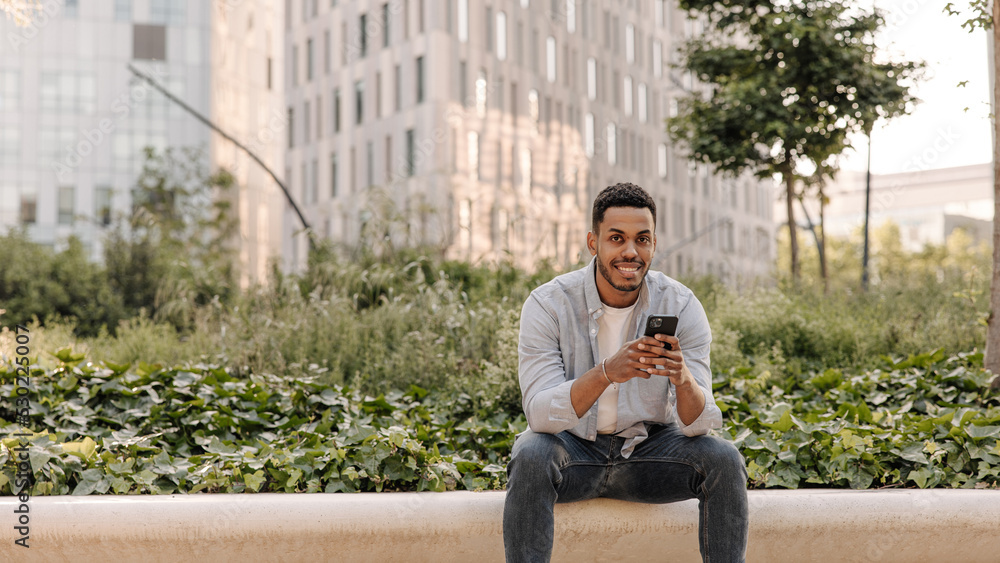 Obraz premium Handsome young african guy with beard walking around city with mobile phone. Brunette man looks at camera sitting outdoors. Technology concept