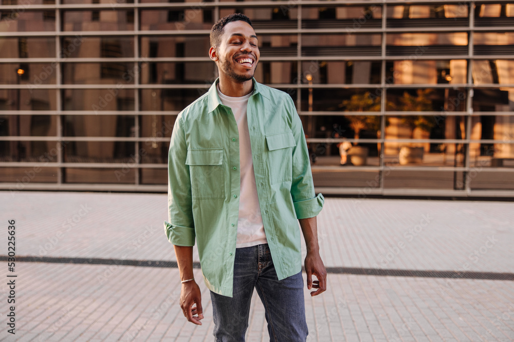 Fototapeta premium Modern young african man with wide smile laughs his eyes closed standing on street. Brunette guy with stubble wears casual clothes. Concept of enjoying moment