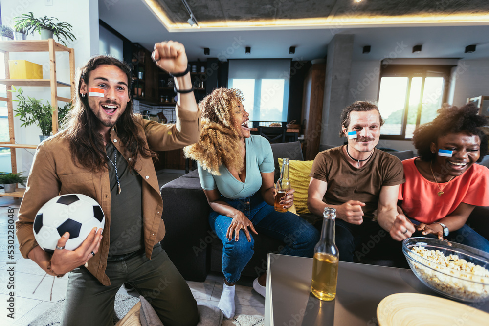 Young group watching sports on television and cheering. Emotional football fans watching football game together at home. Sports Fans with Painted Faces Sitting on a Couch Watch Game on TV