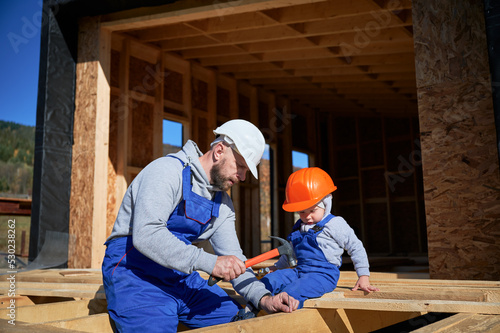 Father with toddler son building wooden frame house in Scandinavian style barnhouse. Boy helping his daddy, hammering nail into plank on construction site on sunny day. Carpentry and family concept.