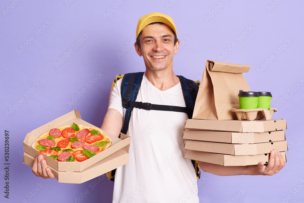 Horizontal shot of satisfied young adult delivery man in yellow cap and T-shirt holding pizza in cardboard flatbox and takeaway coffee isolated on violet background, smiling at camera. Horizontal shot of satisfied young adult delivery man in yellow cap and T-shirt holding pizza in cardboard flatbox and takeaway coffee isolated on violet background, smiling at camera.