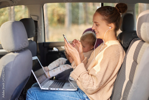 Side view portrait of smiling extremely woman with earphones listening musing from smartphone, working on laptop while sitting with her baby daughter in safety chair on backseat of the auto.