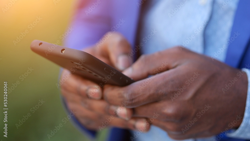 Black man wearing blue blazer hands enter necessary information into modern smartphone. African American person works as entrepreneur outdoors, sunlight