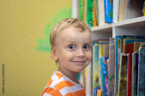 Little blond boy of preschool age smiling in the library. Close-up of a smiling face. Boy with books near the bookcase