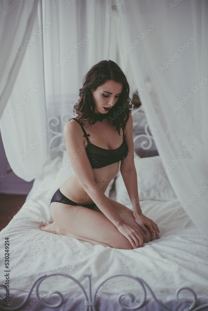 Naklejka premium A young attractive girl with dark hair in black lingerie and a white bathrobe on a white bed in the bedroom. beauty and fashion