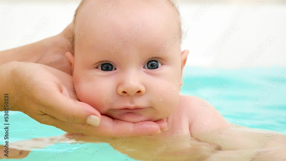 Mother teaches a newborn baby to swim and stay on the water. Two-month-old baby is bathing in a large bathtub. Newborn baby is in the water, bathroom. Hygiene procedures of a newborn baby Mother teaches a newborn baby to swim and stay on the water. Two-month-old baby is bathing in a large bathtub. Newborn baby is in the water, bathroom. Hygiene procedures of a newborn baby
