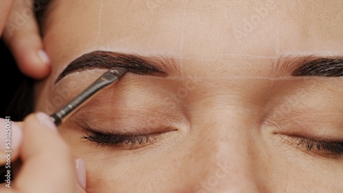 Woman is  on the Eyebrow coloring procedure. Professional lamination procedures of female eyebrows in beauty salon. Closeup view of client face and stylist's hands working.  Beauty care concept. photo