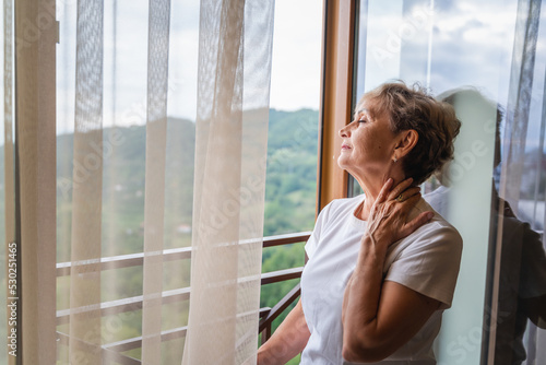 Beautiful happy gray-haired mature woman in a white t-shirt standing by the window enjoying the morning breathing with closed eyes