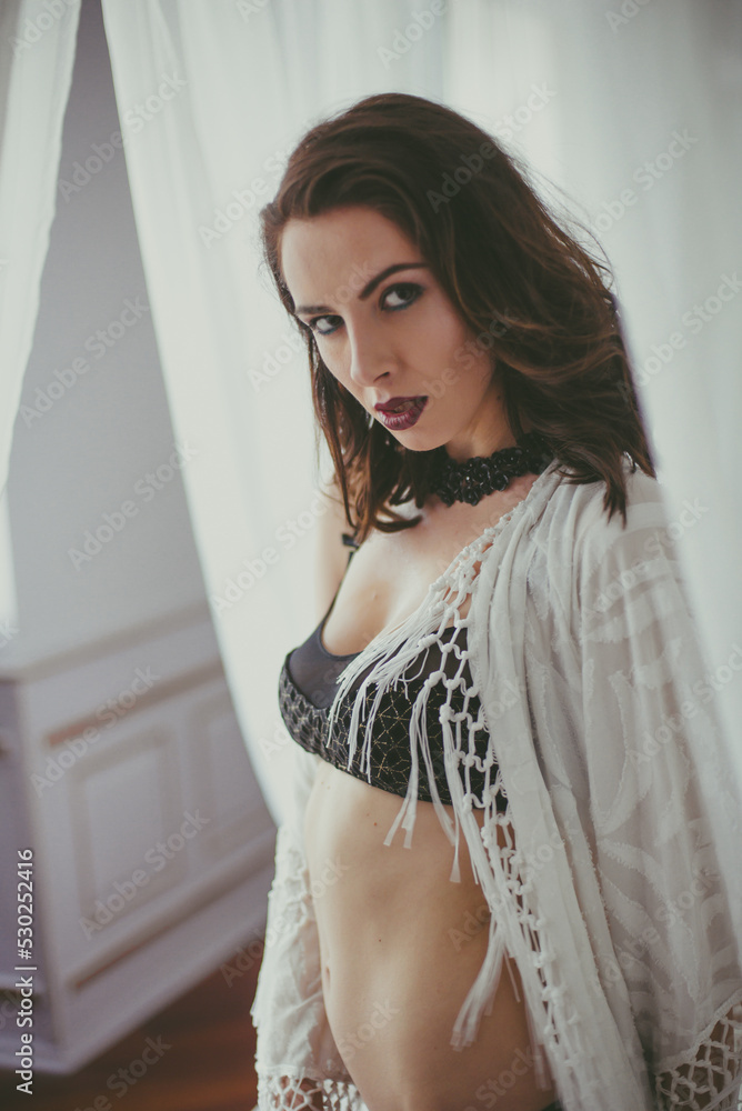 Fototapeta premium A young attractive girl with dark hair in black lingerie with evening make-up and jewelry and a white bathrobe on a white bed in the bedroom. beauty and fashion