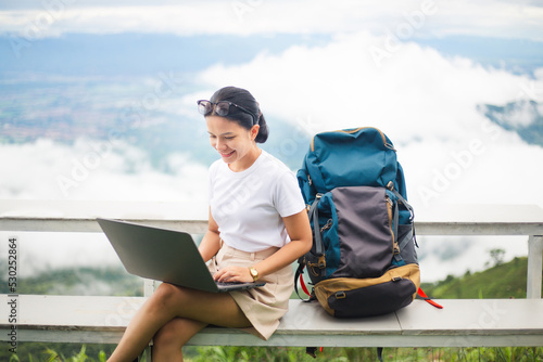Young woman traveler and blogger with backpack using laptop.  Asian female tourist working at computer on vacation trip. Travel and technology concept. Top of mountains, sky and fog background.