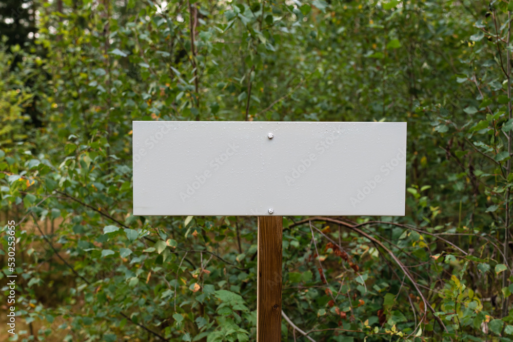 Blank white sign in nature with water droplets on the surface and green leaves in the background