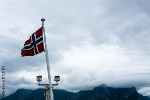 Norwegian flag waving on board a ship in Norwegian sea, Atlantic ocean with cloud covered mountains in the background.